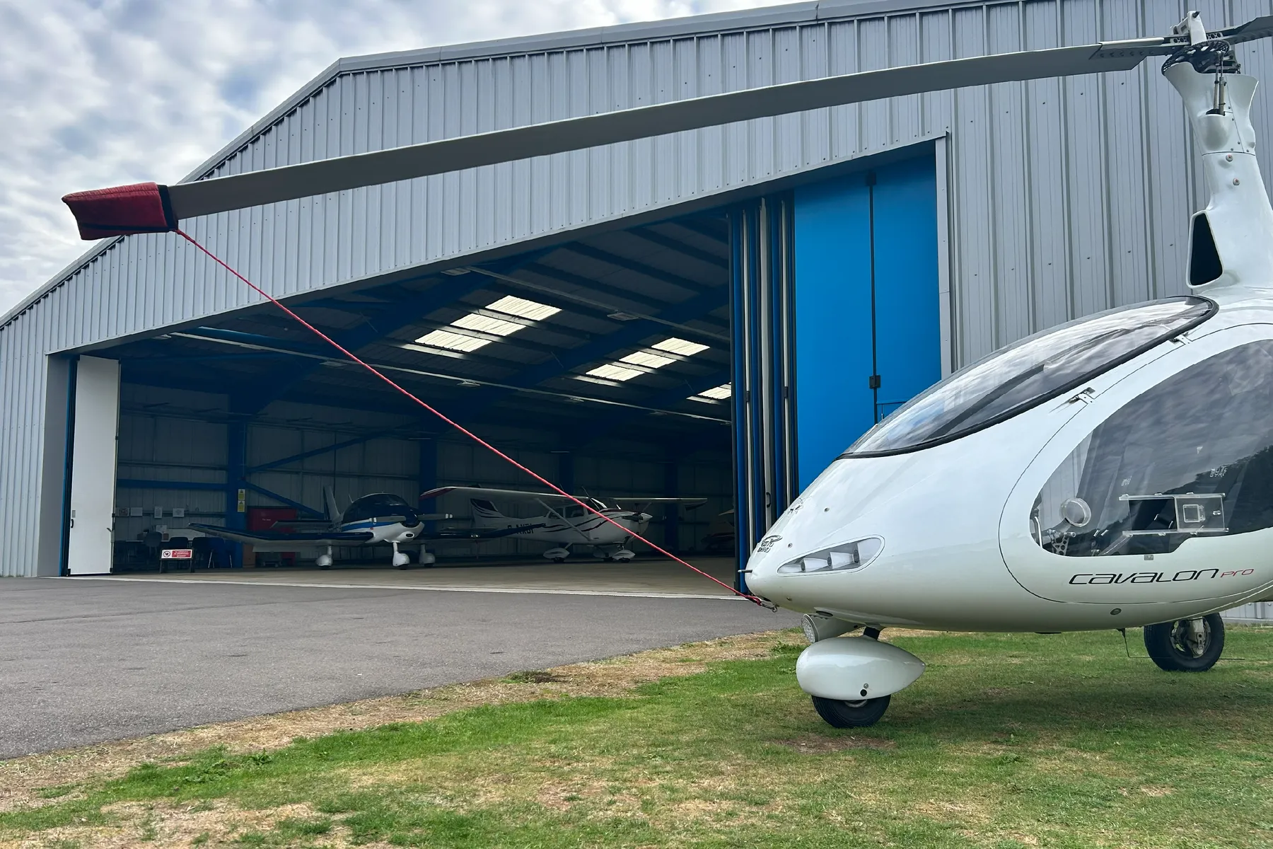A gyroplane parked on the grass in front of an open hangar with light aircraft inside